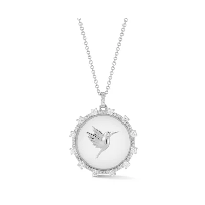 The Floating Hummingbird Necklace is a silver piece with a round pendant, engraved with a hummingbird at the center and bordered by small, sparkling stones.