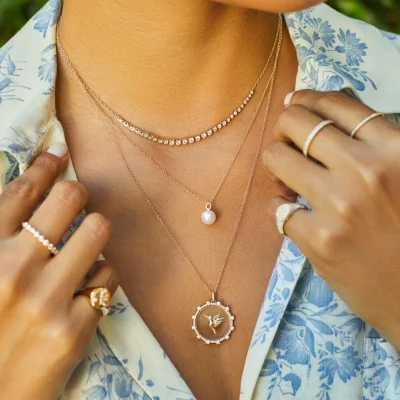 EF-Spring_24_Hummingbird-Pearl_01915_GabrielSweet_Photography_1488x1488 A person wearing the Floating Hummingbird Necklace layered with other gold necklaces, several gold rings, and a light blue floral shirt.