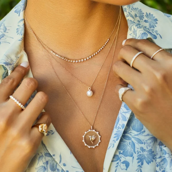 A person wearing the Floating Hummingbird Necklace layered with other gold necklaces, several gold rings, and a light blue floral shirt.