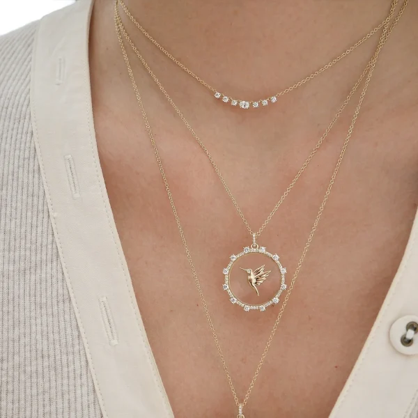 A close-up of a person wearing the Floating Hummingbird Necklace—three layered gold chains, each with small diamonds and a circular pendant featuring a hummingbird, over a cream-colored buttoned shirt.
